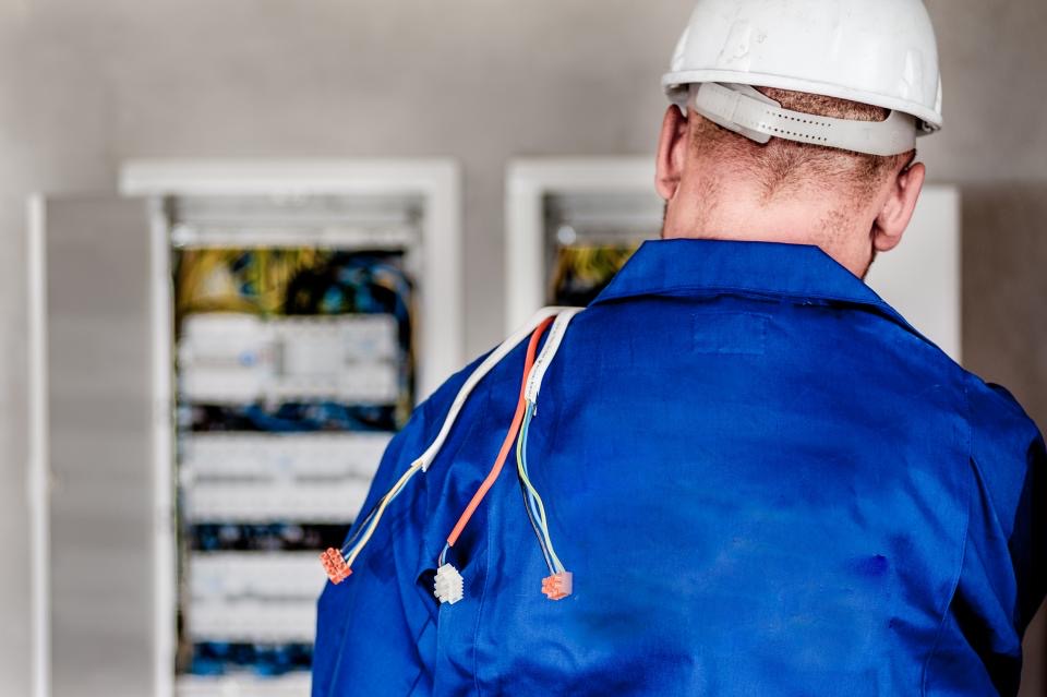 Electrician in a blue work jacket and hard hat at an open electrical panel